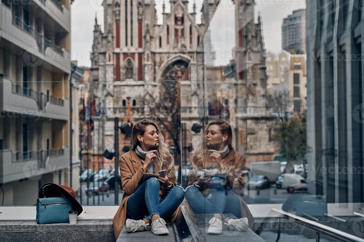 disfrutando de la música favorita. atractiva joven escuchando música usando su teléfono inteligente y mirando su reflejo en la caja de vidrio mientras pasa tiempo sin preocupaciones en la ciudad foto