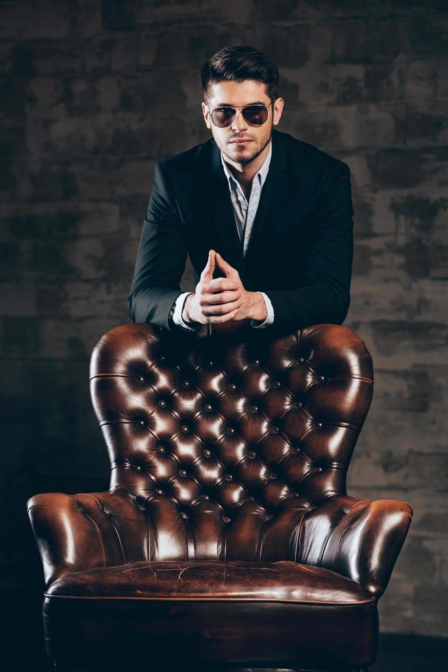 Charming and elegant. Young handsome man in suit and sunglasses leaning on leather chair and looking at camera while standing against dark grey background photo