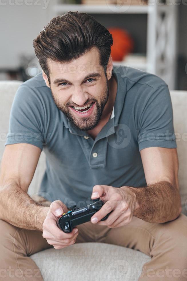 Games always fun. Cheerful young handsome man playing video game and looking at camera while sitting on the couch at home photo