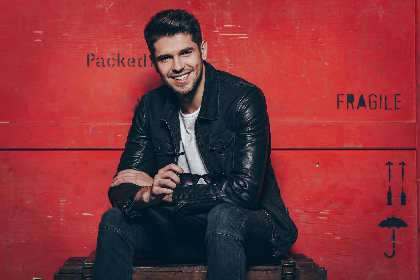 Trendy and charming. Handsome cheerful young man holding his sunglasses and looking at camera with smile while sitting on wooden chest against red background photo