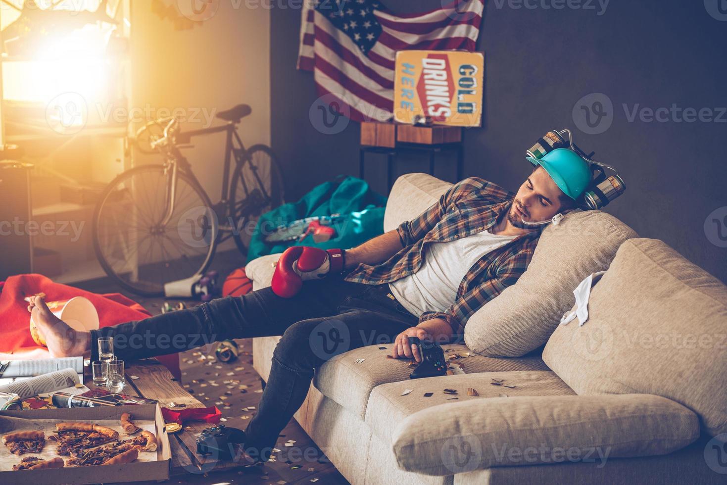 Desmayado. Un joven apuesto con guante de boxeo y sombrero de cerveza tomando una siesta en un sofá en una habitación desordenada después de la fiesta foto