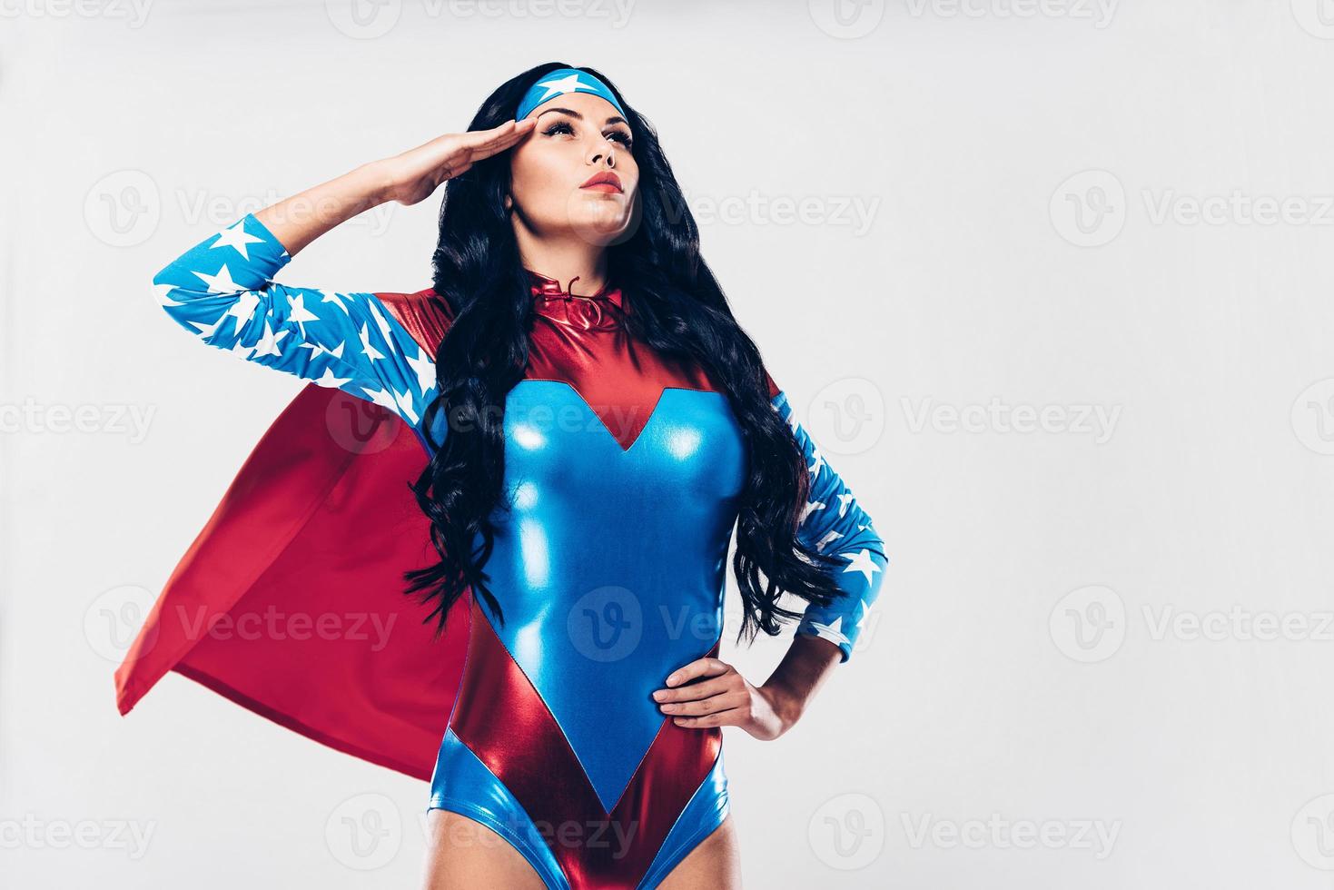 On duty with fighting crime. Low angle view of beautiful young woman in superhero costume looking away and holding hand near head while standing against white background photo