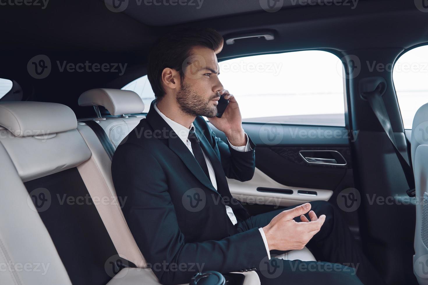 Always available. Handsome young man in full suit talking on the smart phone while sitting in the car photo