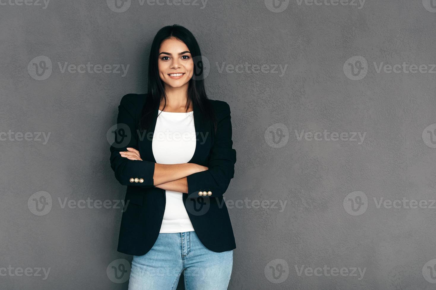 segura y hermosa. mujer joven atractiva con ropa informal elegante que mantiene los brazos cruzados y mira la cámara con una sonrisa mientras se enfrenta a un fondo gris foto