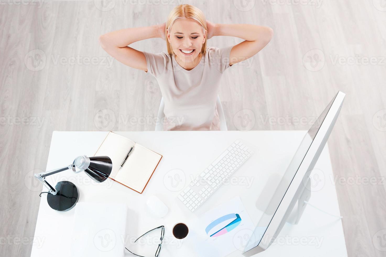 Taking a minute break. Top view of beautiful young smiling woman holding hands behind head while sitting at her working place photo