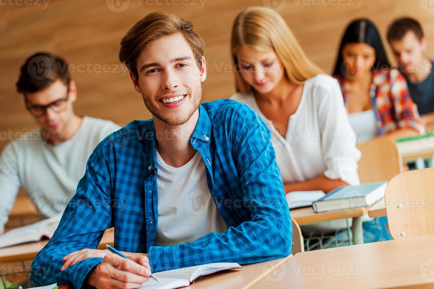 Showing a dedication to education. Handsome young man writing in note pad and looking at camera while sitting at front desk in the classroom photo