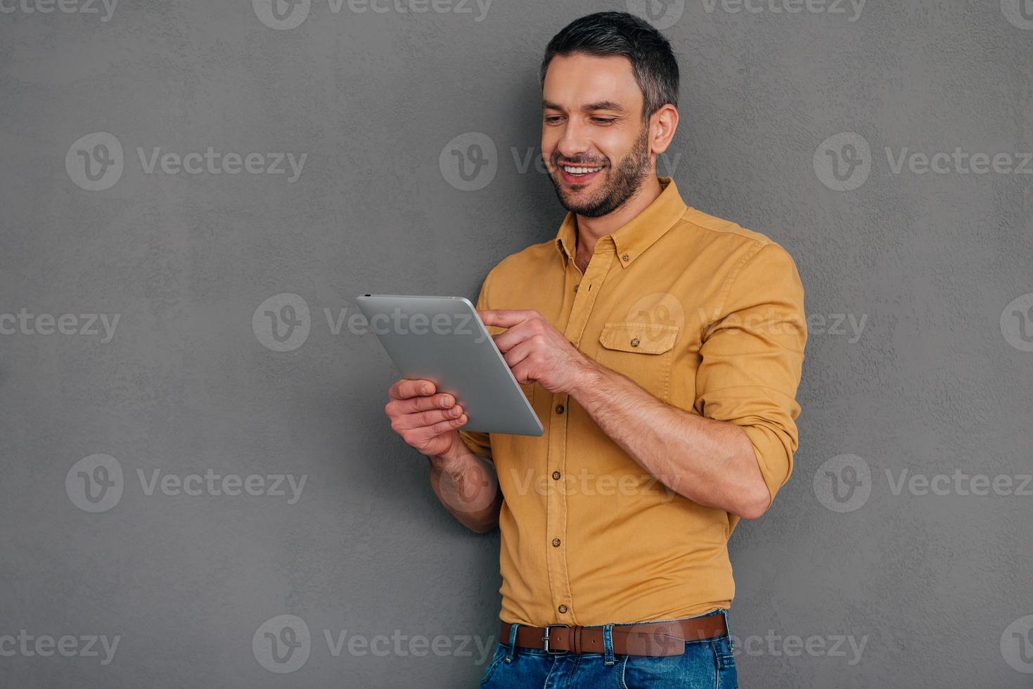 Examining his brand new tab. Handsome mature man holding digital tablet and looking at it with smile while standing against grey background photo