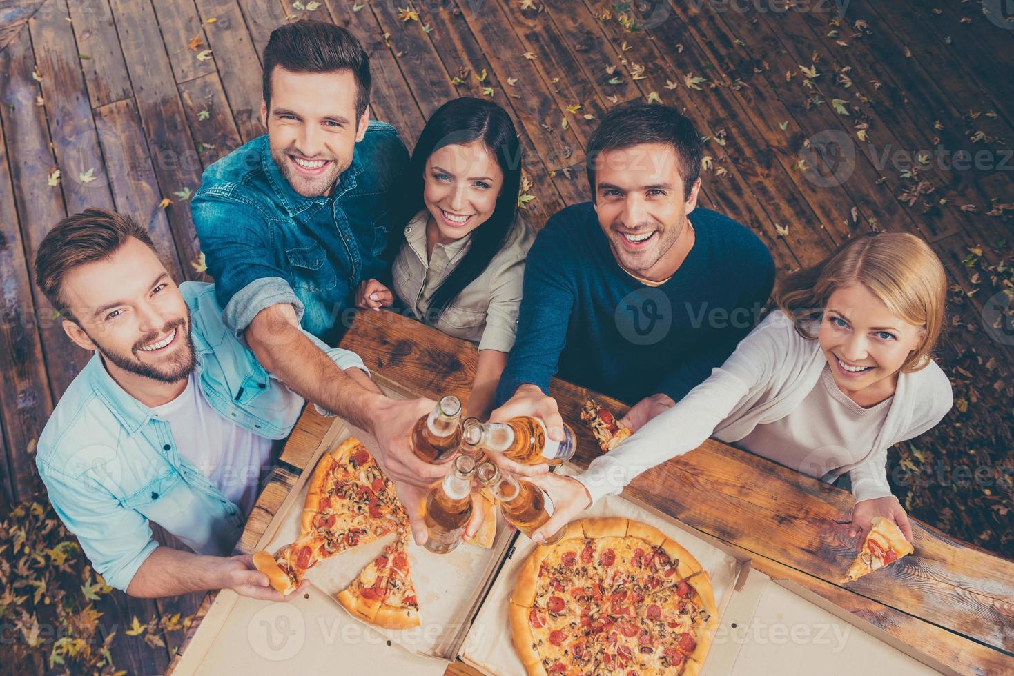 celebrando ese día. vista superior de cinco jóvenes felices tintineando vasos con cerveza y mirando a la cámara mientras están de pie al aire libre foto