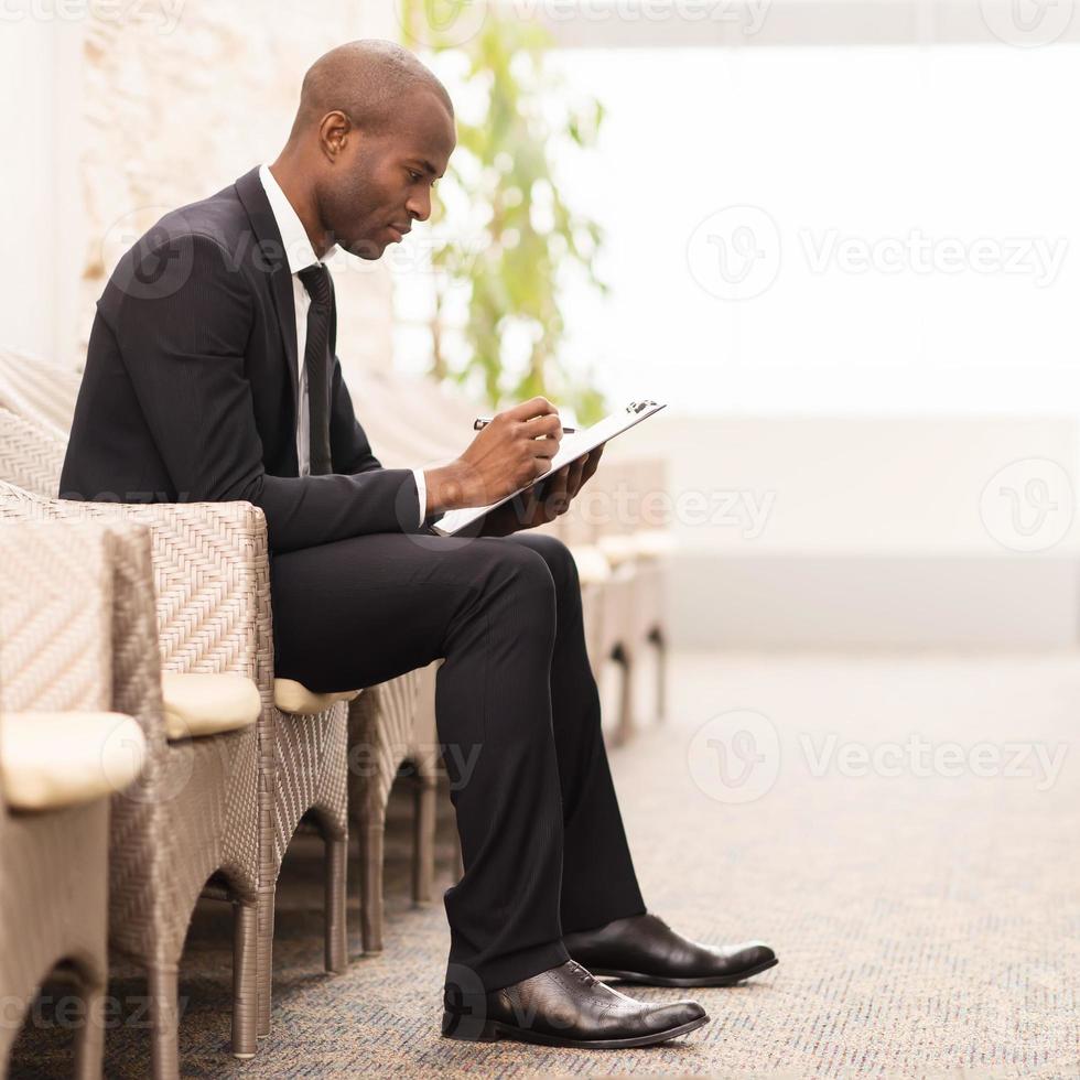 Making notes. Side view of confident young African businessman writing something in his note pad while sitting on the chair photo