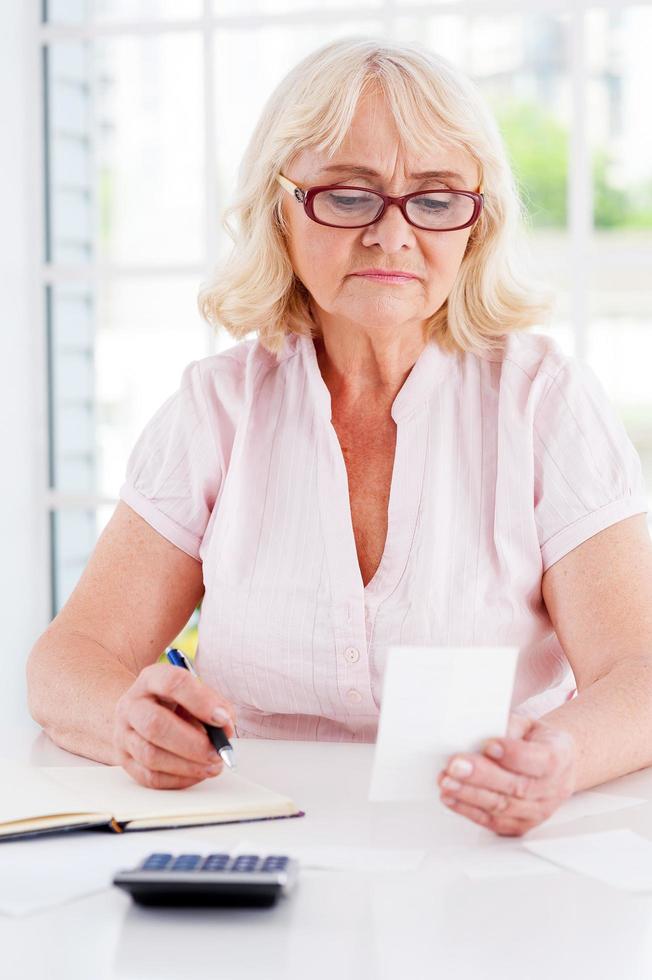 calculo de gastos. anciana concentrada sosteniendo un billete y escribiendo algo en su bloc de notas mientras estaba sentada en la mesa foto