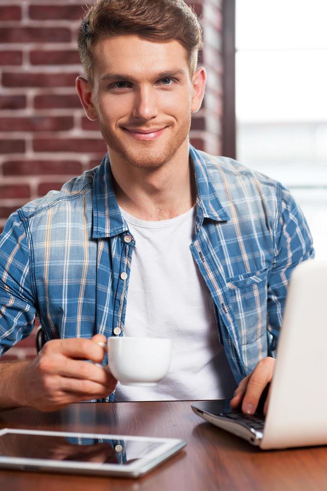 Coffee time. Handsome young man holding coffee cup and smiling while sitting at his working place photo