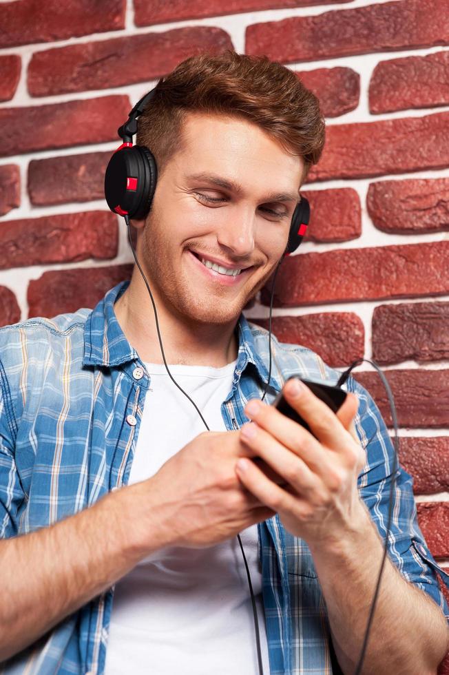 Listening to his favorite music. Handsome young man listening to the MP3 Player while standing against brick wall photo