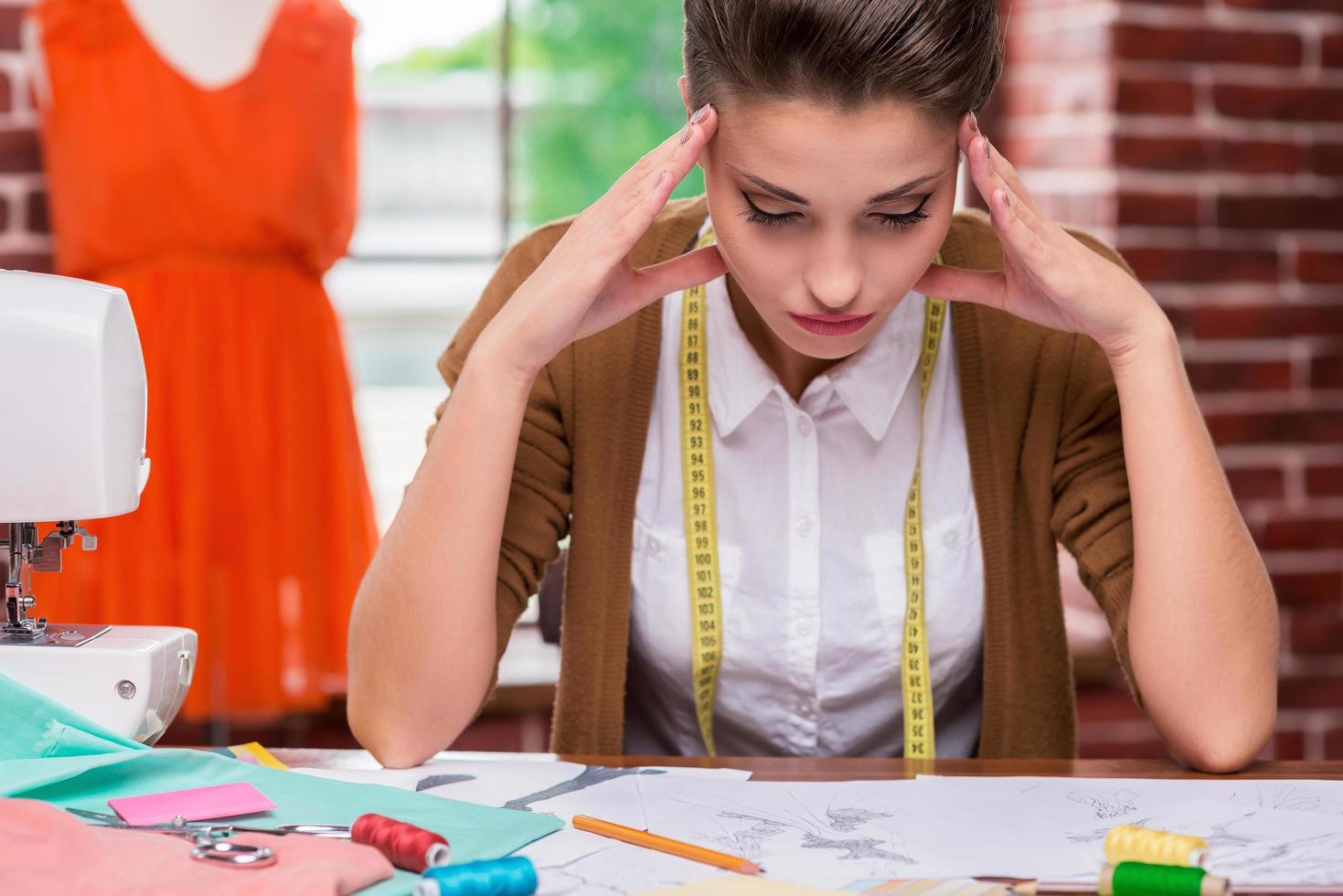 I need some fresh ideas. Tired young woman holding head in hands and looking at fashion sketches while sitting at her working place photo