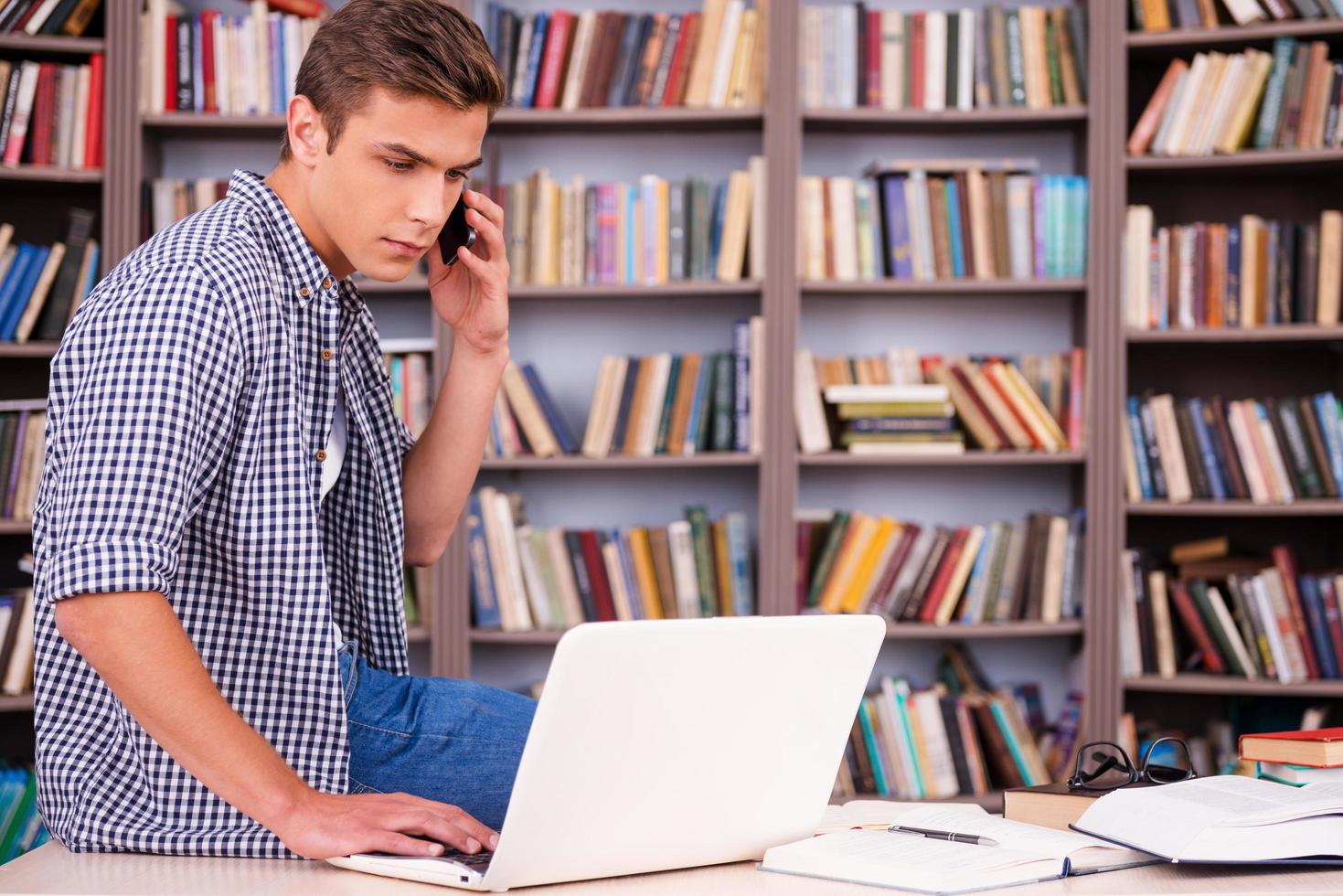 Full concentration on studying. Confident young man working on laptop and talking on the mobile phone while leaning at the desk and in font of bookshelf photo