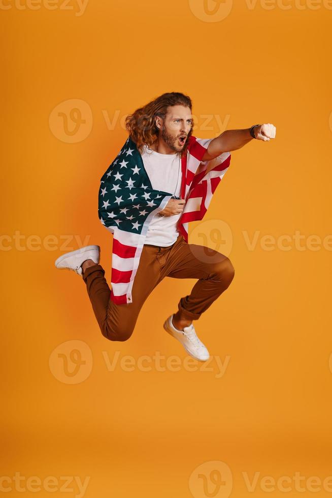 Confident young man in casual clothing making a face and punching while hovering covered in American flag against yellow background photo