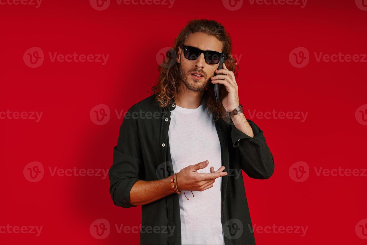 joven ocupado con ropa informal mirando a la cámara y hablando por teléfono mientras se enfrenta a un fondo rojo foto