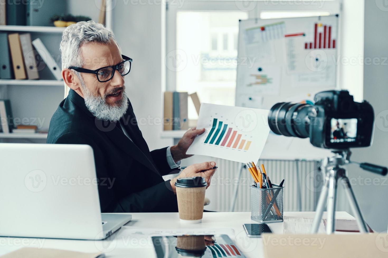 Confident mature man in elegant suit showing chart and sharing business experience while making social media video photo