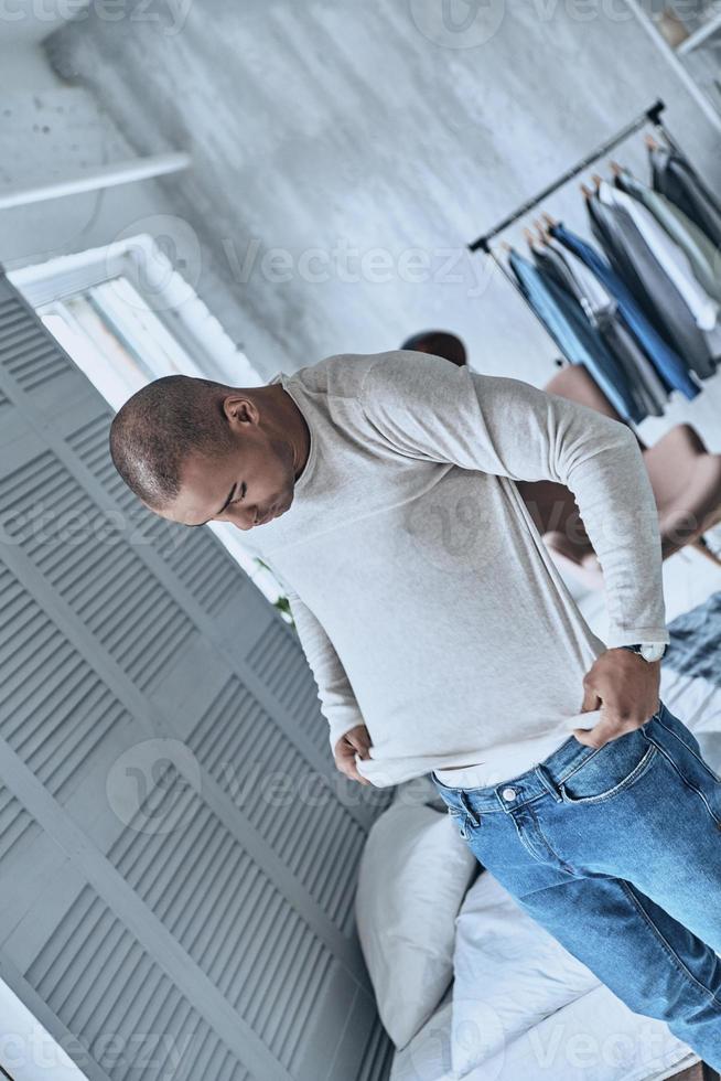 Finally ready. Top view of handsome young African man putting on shirt while standing in the bedroom at home photo