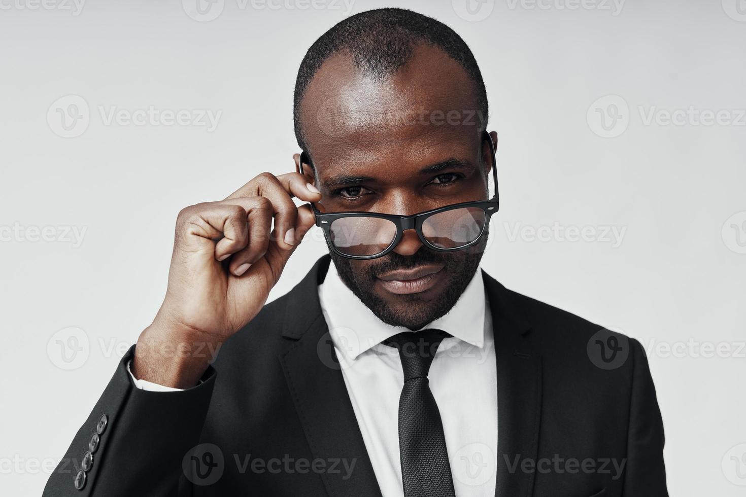 joven africano confiado en ropa formal mirando a la cámara y ajustando los anteojos mientras está de pie contra el fondo gris foto