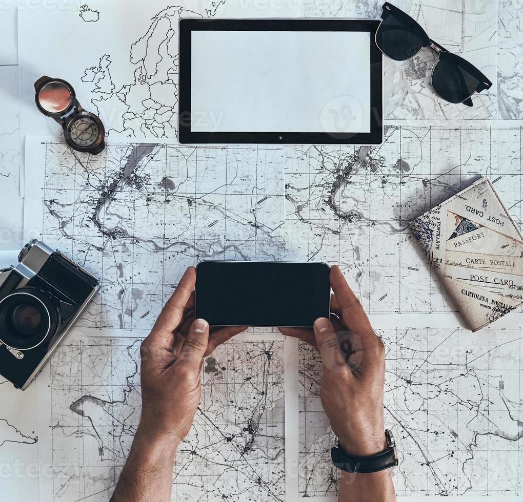 New adventure. Close up top view of man using smart phone with sunglasses, photo camera, compass and passport lying on map around