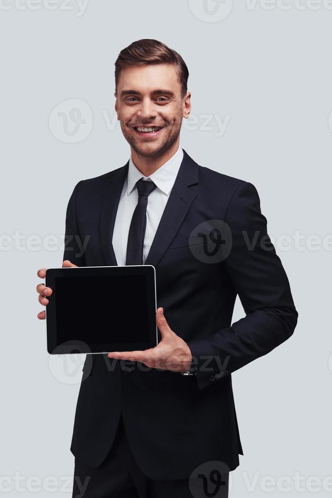 Using modern technologies. Good looking young man in full suit holding digital tablet and looking at camera while standing against grey background photo