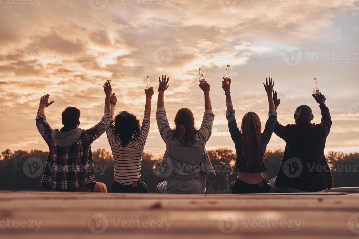 amistad que dura para siempre. vista trasera de jóvenes con ropa informal brindando con botellas de cerveza mientras se sientan en el muelle foto