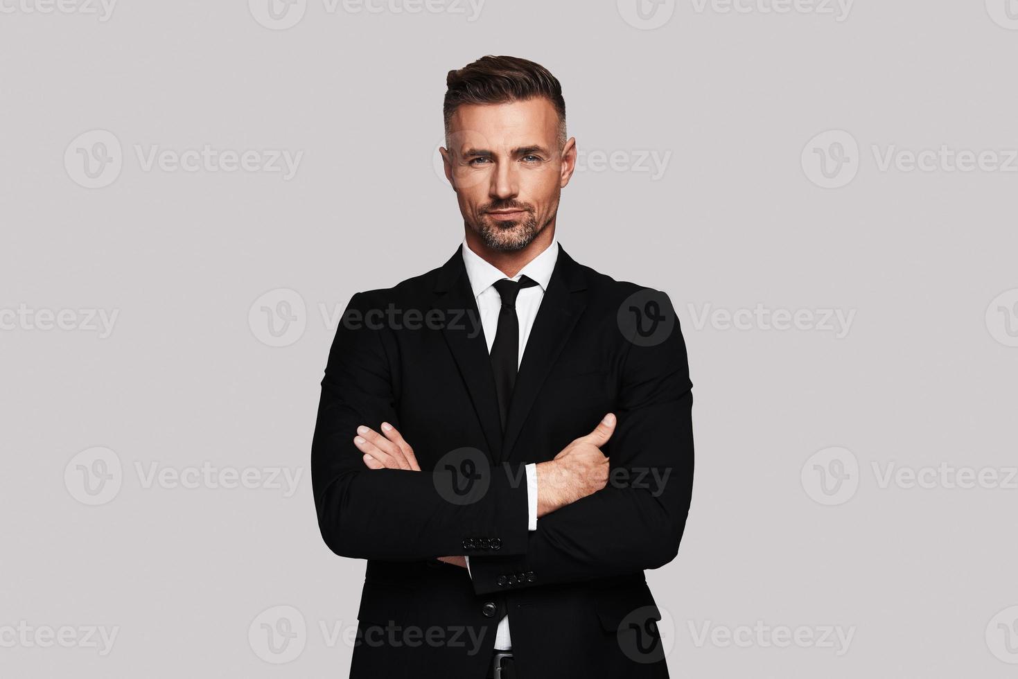 Business expert. Handsome young man in full suit keeping arms crossed and looking at camera while standing against grey background photo