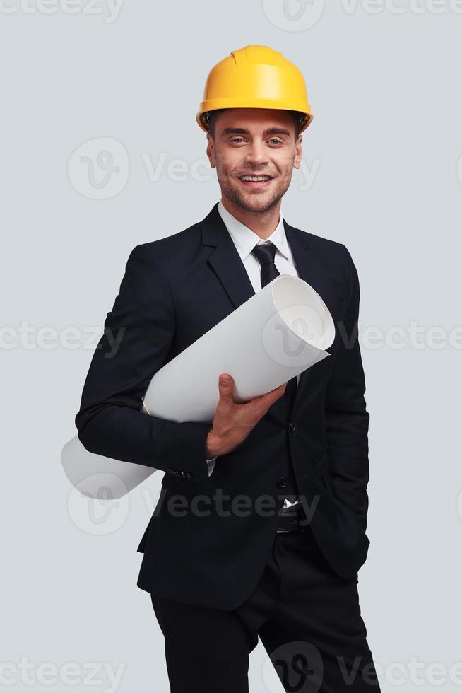 Young architect. Good looking young man in hardhat carrying blueprint and smiling while standing against grey background photo