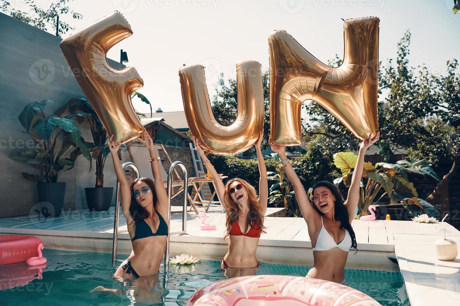 Attractive young women in swimwear smiling and lifting up balloons while standing in the pool outdoors photo