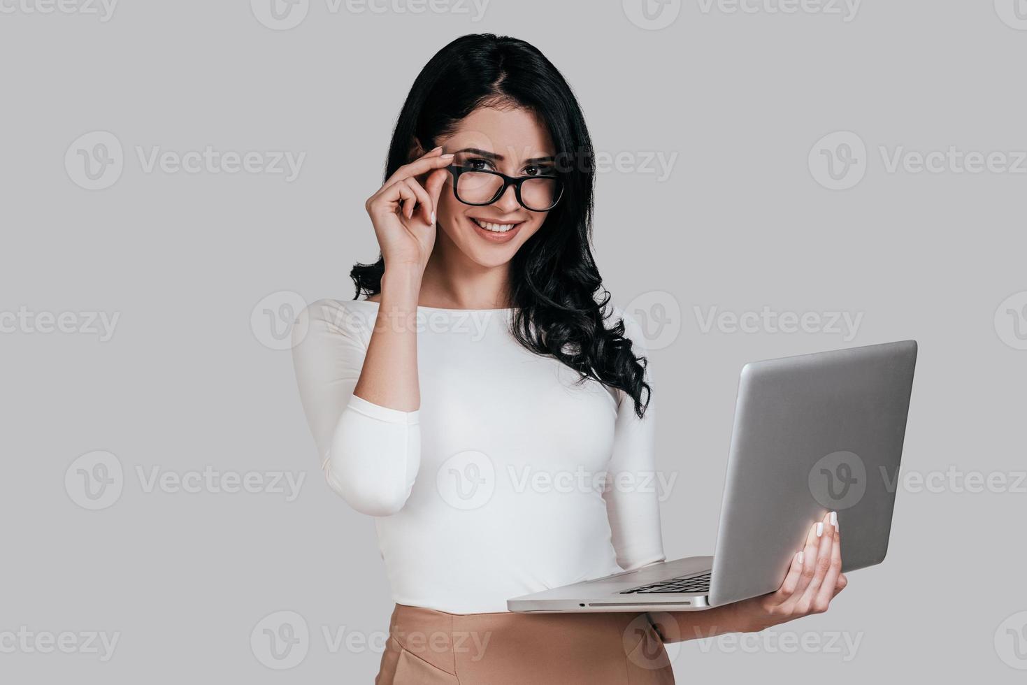 mujer joven hermosa en ropa casual inteligente sosteniendo una computadora portátil y ajustando sus anteojos mientras está de pie contra un fondo gris foto