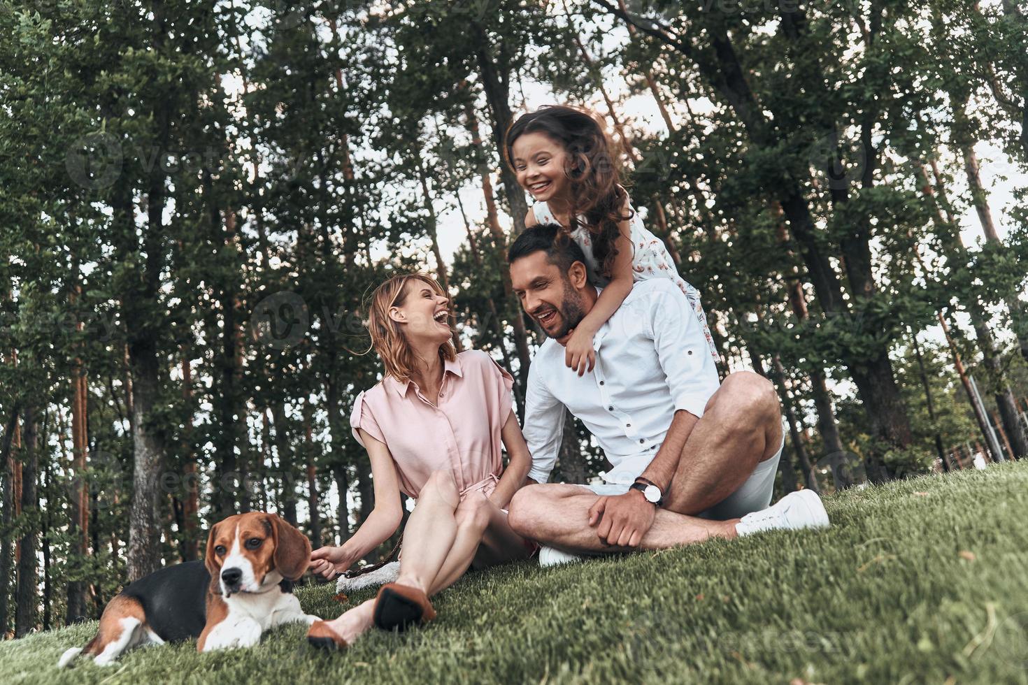Enjoying every moment together. Happy young family of three with dog smiling while sitting on grass in park photo