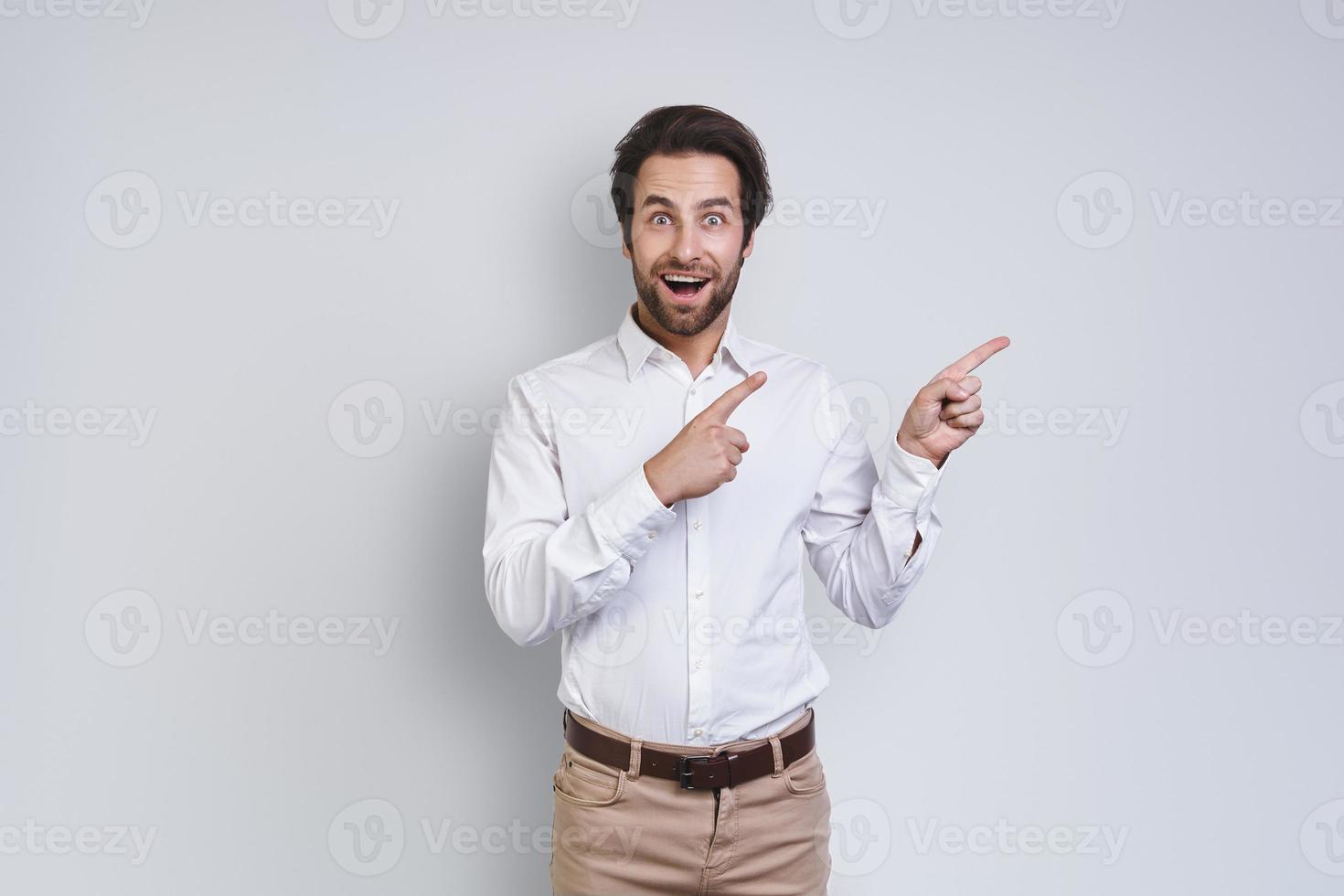 Handsome young smiling man in white shirt looking at camera and pointing away while standing against gray background photo