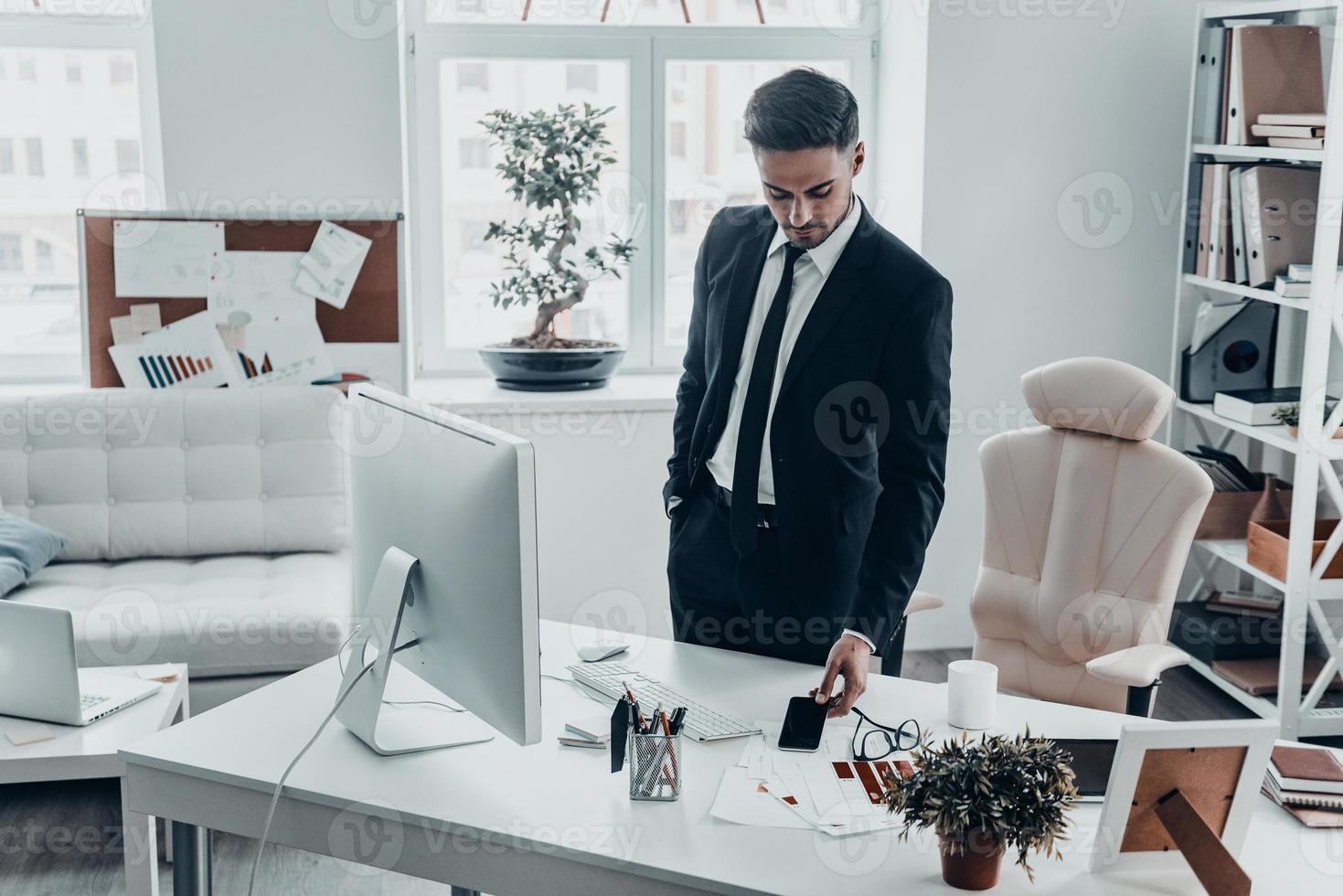 No more phone calls for today. Good looking young man in full suit laying his smart phone on the desk while standing in the office photo