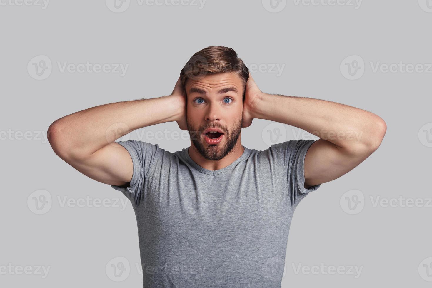 Totally lost. Shocked young man covering ears with hands and looking at camera while standing against grey background photo