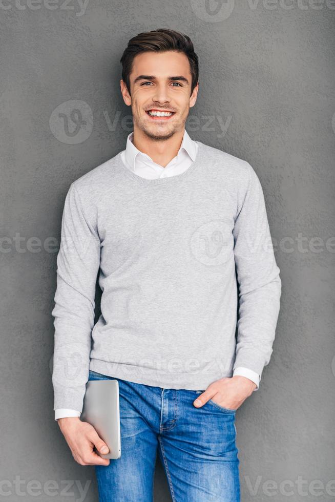 Mr. Expertise. Confident young man looking at camera with smile and carrying digital tablet while standing against grey background photo