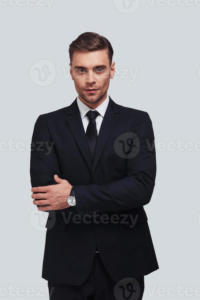 Confident in his style. Handsome young man in full suit keeping arms crossed and looking at camera while standing against grey background photo