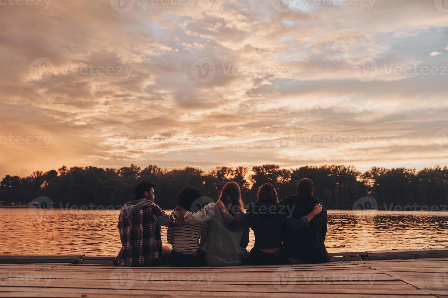 vida llena de amistad. vista trasera de jóvenes con ropa informal abrazándose mientras están sentados en el muelle foto