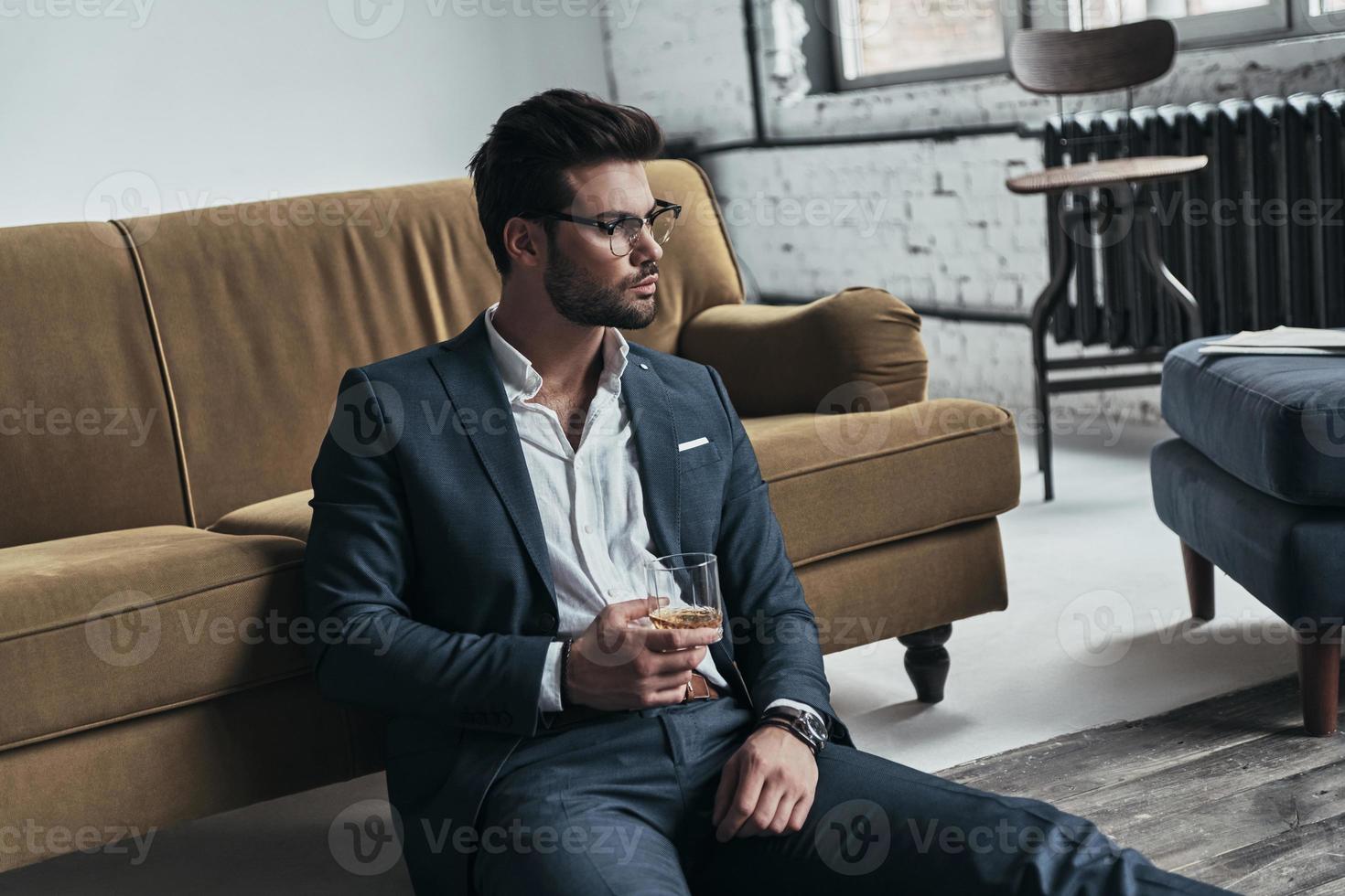 Enjoying favorite drink. Handsome young man in full suit holding a drinking glass and looking away while sitting on the floor at home photo
