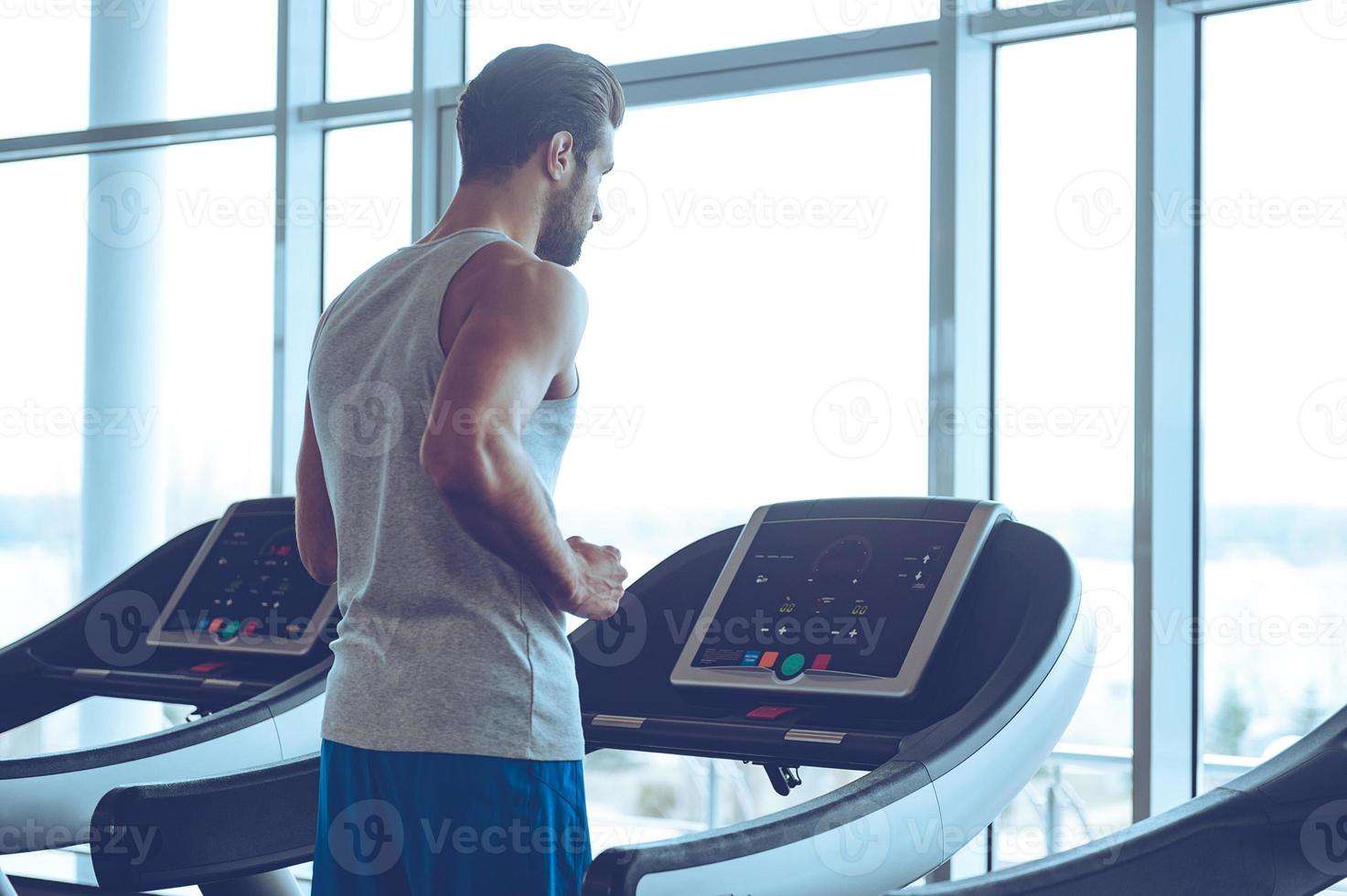 Body power. Young man in sportswear running on treadmill in front of window at gym photo