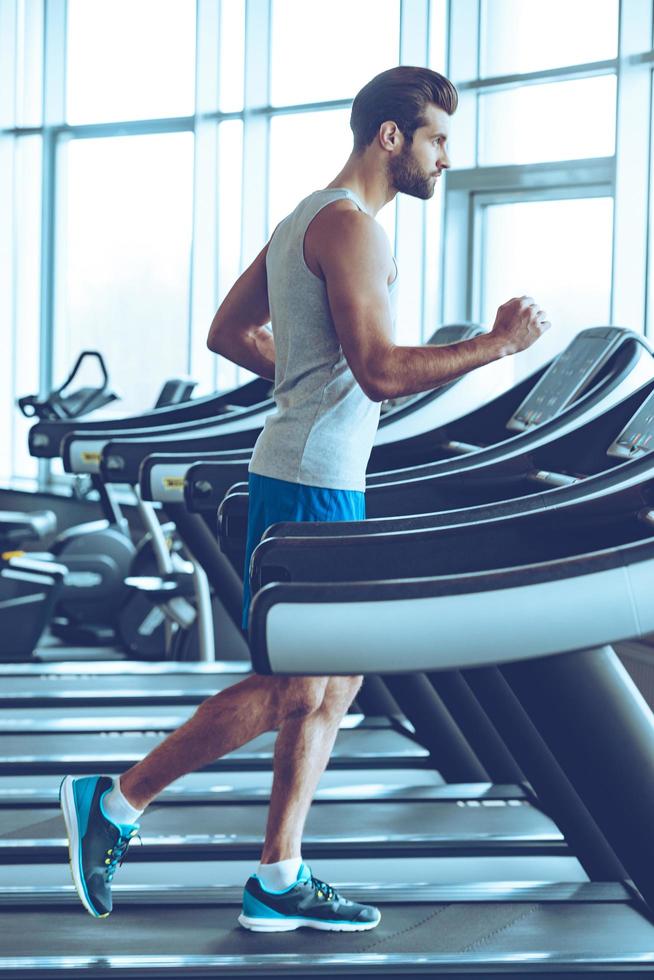 Jogging on treadmill. Side view full length of young man in sportswear running on treadmill at gym photo