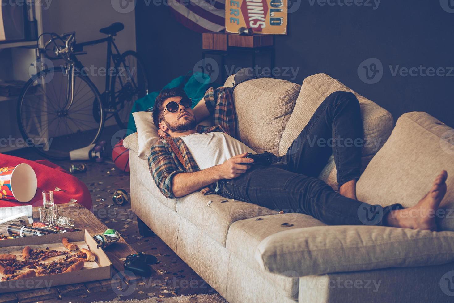 Party is over. Young handsome man in sunglasses lying down on sofa with joystick in his hand in messy room after party photo