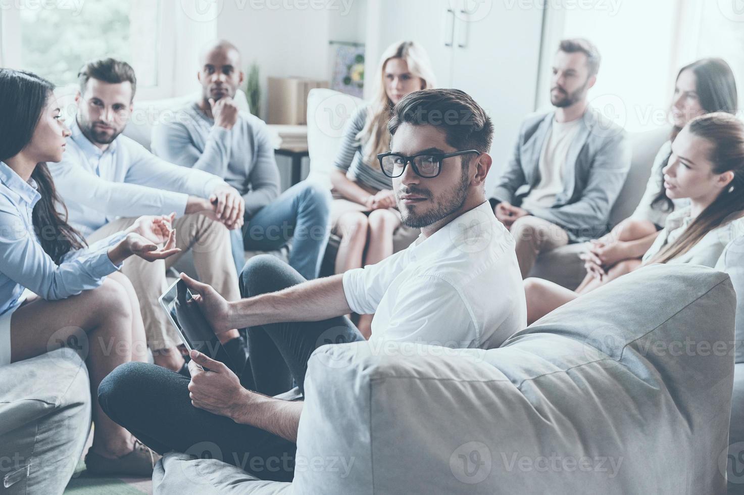 Confident life coach. Group of young people sitting in circle and discussing something while young man holding digital tablet and looking over shoulder photo