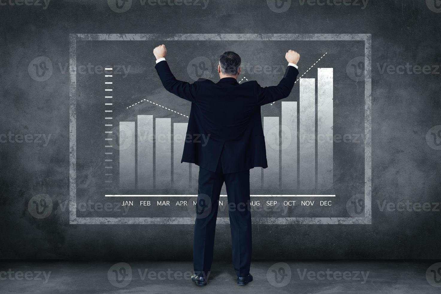 When business goes well. Rear view full length of mature businessman holding arms outstretched while standing against grey background with illustration of chart photo