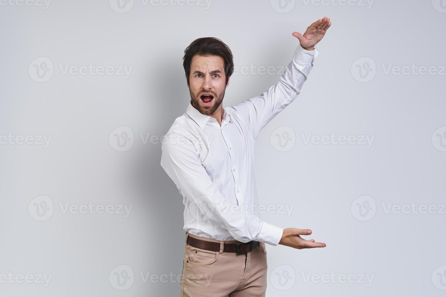 Surprised young man in white shirt looking at camera and gesturing while standing against gray background photo