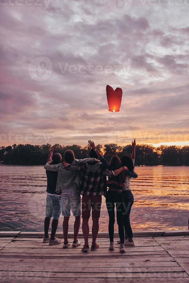Flying away. Full length rear view of young people embracing and gesturing while looking at floating sky lantern photo