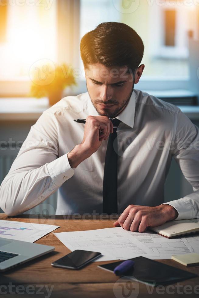 Examining new contracts. Pensive young handsome man looking at documents and keeping hand on chin while sitting at his working place photo