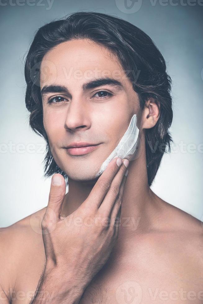 Fresh and clean shaving. Portrait of handsome young shirtless man applying cream on face and looking at camera while standing against grey background photo