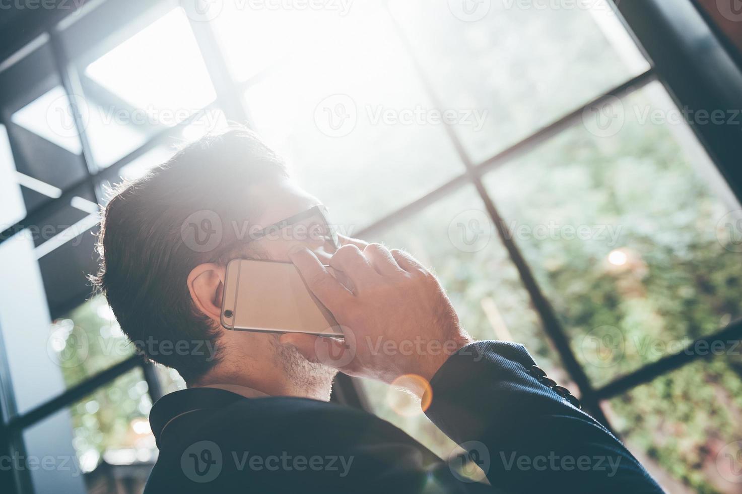 Man on the phone. Low angle view of young man talking on the mobile phone and looking away while standing in front of the big window indoors photo