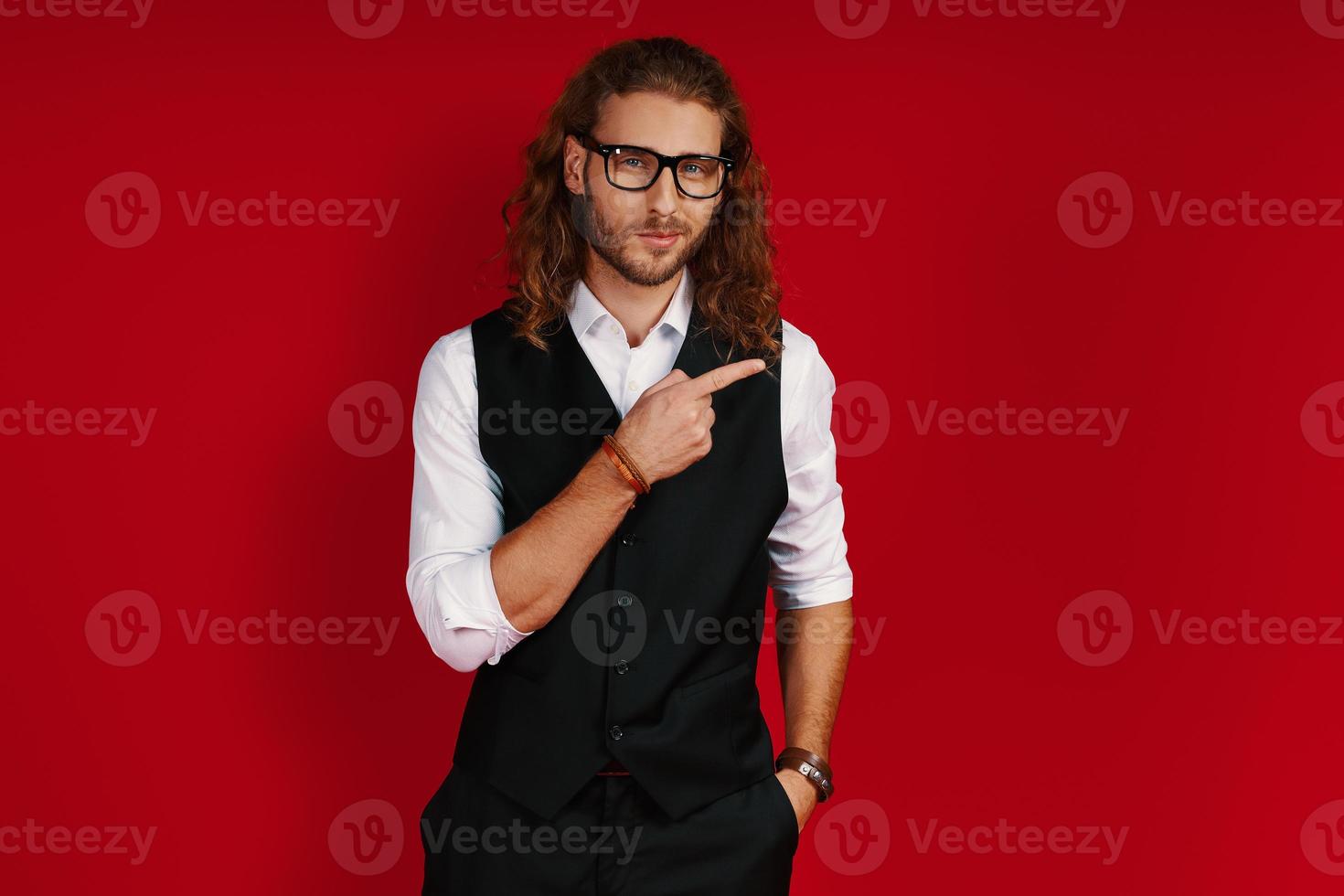 Happy young man in elegant clothing looking at camera and pointing copy space while standing against red background photo