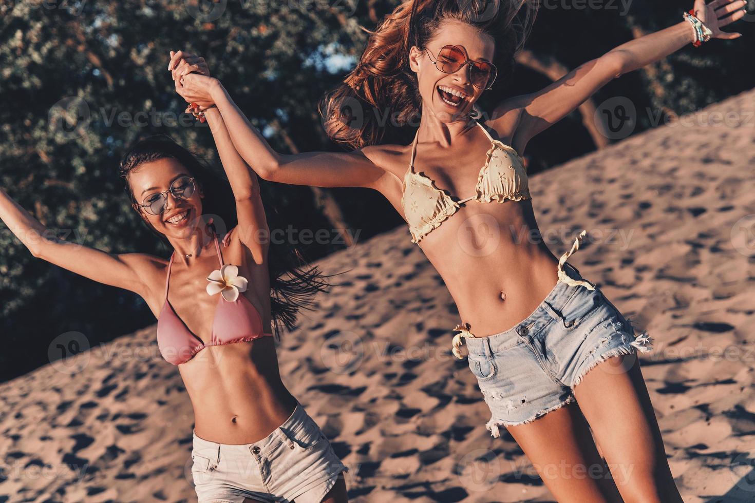 mejores amigos tienen la mejor de la diversión. dos mujeres jóvenes atractivas en pantalones cortos y trajes de baño sonriendo y tomándose de la mano mientras corren en la playa foto
